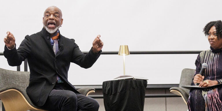 A nearly bald man with a white mustache and goatee, wearing a colorful scarf and black suit jacket, gestures with his hands while sitting to the left of a small table with a gold lamp. A woman with black, curly hair wearing a black dress with a colored streak pattern, sits to the right and holds a microphone with a folder in her lap.