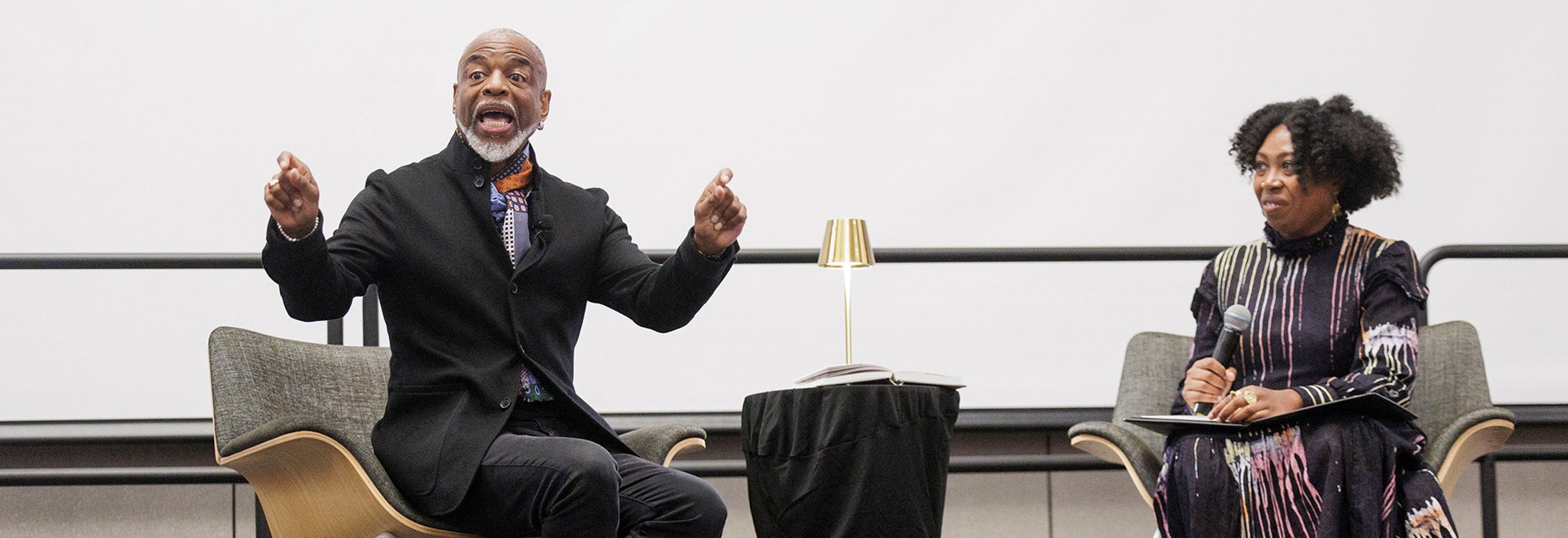 A nearly bald man with a white mustache and goatee, wearing a colorful scarf and black suit jacket, gestures with his hands while sitting to the left of a small table with a gold lamp. A woman with black, curly hair wearing a black dress with a colored streak pattern, sits to the right and holds a microphone with a folder in her lap.