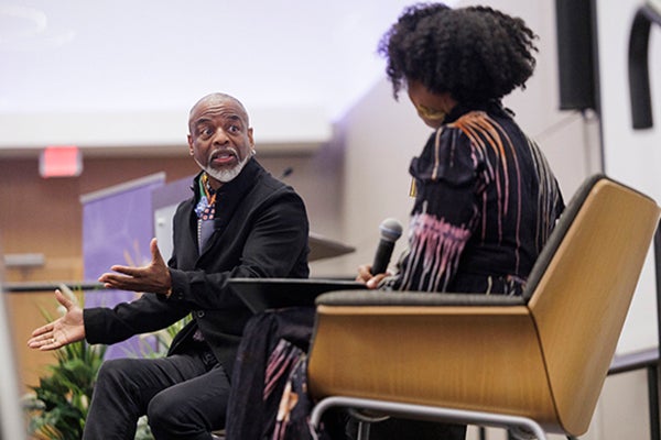 A nearly bald man with a white mustache and goatee, wearing a colorful scarf and black suit jacket, gestures with his hands while sitting to the left of a small table with a gold lamp. A woman with black, curly hair wearing a black dress with a colored streak pattern sits to the right and holds a microphone.