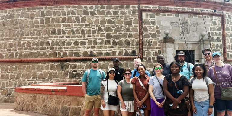 Alt Text description, no acronyms: A group of young men and women stand outside of a brick fortress.