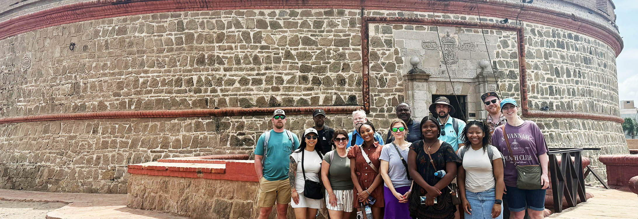 Alt Text description, no acronyms: A group of young men and women stand outside of a brick fortress.