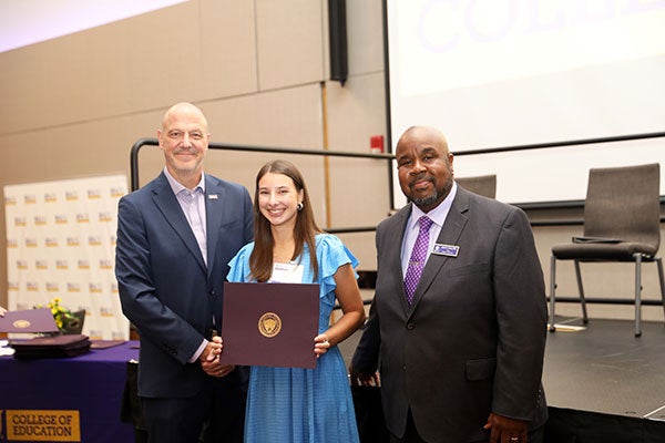 A brunette ECU College of Education student, Lexi Lozner, wears a blue dress and holds an award as she poses with ECU Vice Chancellor for University Advancement, Christopher Dyba, and ECU College of Education’s Dean Andre Green. Both men are wearing purple button-up shirts and dark suits.