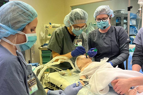 Four Pirate nurses, wearing gray surgical scrubs and blue masks, surround a gurney holding a medical mannequin.