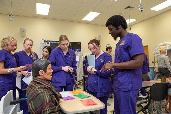 Six Pirate nurses wearing purple surgical scrubs stand around a seated medical mannequin in a simulation of patient care.