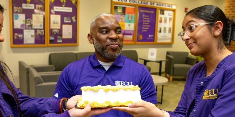 Two women, dressed in purple scrubs, hold the side of a cake beneath the face of a man seated between them.