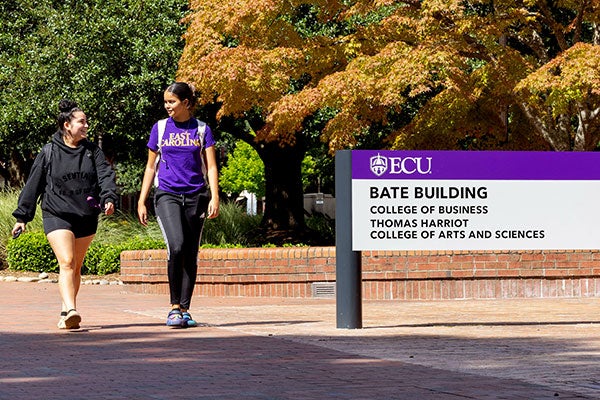 East Carolina University student Lauren Aponte, left, wears a black sweatshirt and black shorts with a backpack on her shoulders while smiling and talking with Lynoshka Padilla, who is wearing a purple T-shirt with East Carolina spelled in gold along with black athletic pants and a backpack. Padilla smiles back at Aponte as they look at each other while walking past the Bate Building on campus.  