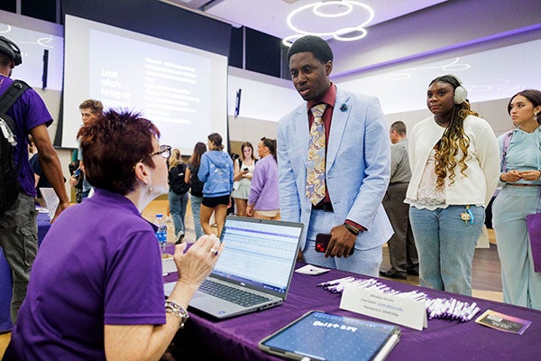 A student in a blue suit speaks to a woman sitting behind a table at a job fair.