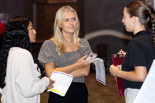 A young woman with blonde hair speaks to a woman with a ponytail wearing a dark shirt while another young woman looks on. 