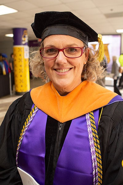 A woman with light hair and glasses wearing academic regalia poses for a photo indoors.