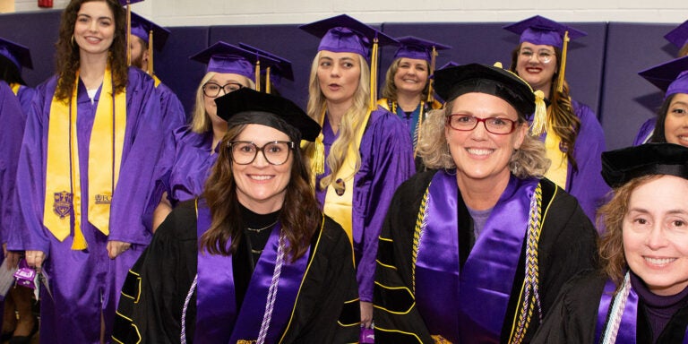 Three women in black graduation regalia pose with a group of women and men behind them in purple graduation attire.