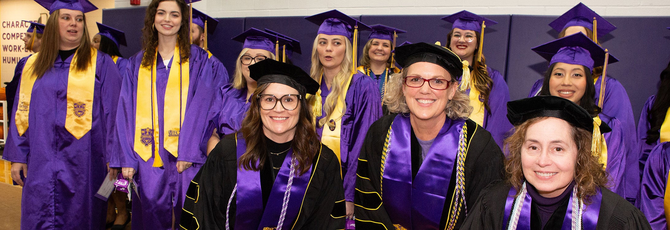 Three women in black graduation regalia pose with a group of women and men behind them in purple graduation attire.