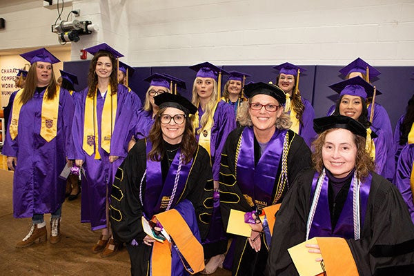 : Three women in black graduation regalia pose with a group of women and men behind them in purple graduation attire. 