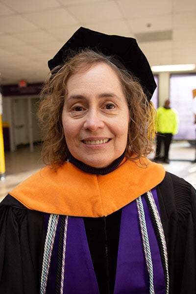 A woman with short brown hair wearing academic regalia poses for a photo indoors.