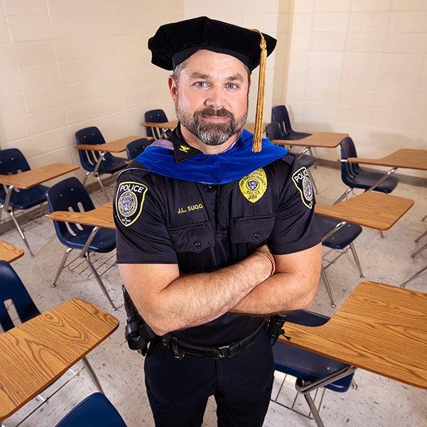 A man in a police uniform and a graduation cap with gold tassel stands with arms crossed in a classroom with desks.