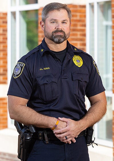 A man in a police uniform stands with his hands crossed on his belt in front of a red brick building.