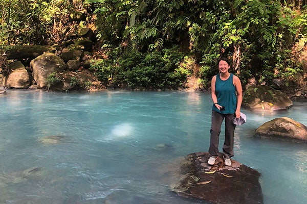 A woman stands on a rock by a turquoise river surrounded by lush green vegetation and large stones.