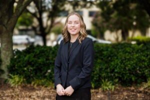 Rebecca Cloninger, wearing a jacket and dark dress, stands on the lawn in front of the Brody School of Medicine.