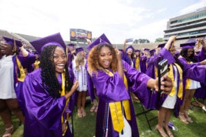 Two women who are surrounded by many new graduates wearing purple gowns and mortarboards smile while taking a selfie at East Carolina University’s commencement.