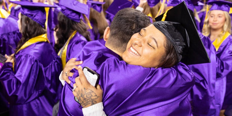 A woman wearing a purple graduation cap and gown holding a phone hugs a man in a graduation gown.