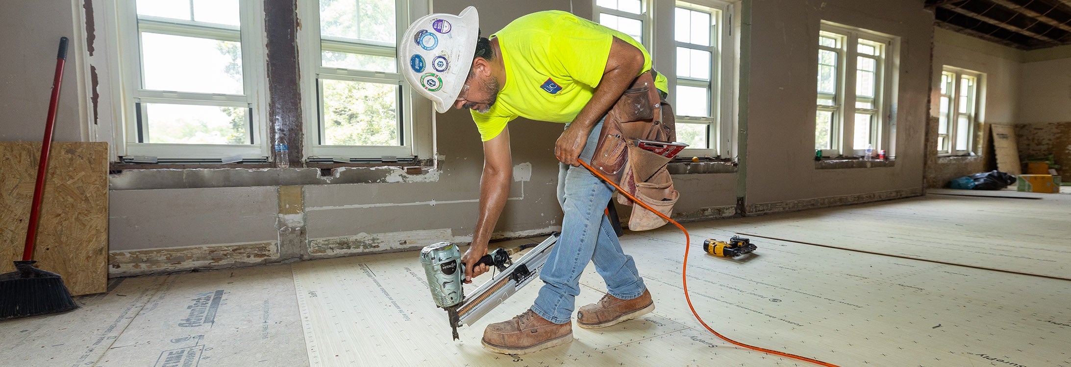 A man in a neon yellow construction shirt and white hard hat holds a nail gun to the floor of the Whichard building on campus.