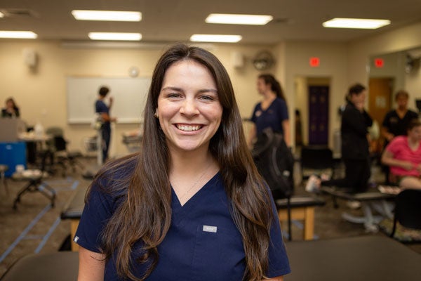 A woman with long dark hair wearing blue medical scrubs smiles as people in the background talk and work together on occupational therapy tasks.