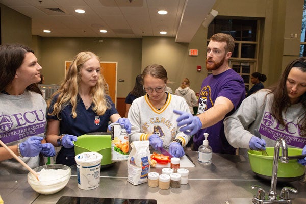 A bearded man in a purple t-shirt talks with a group of women who are preparing food in a large conference room.