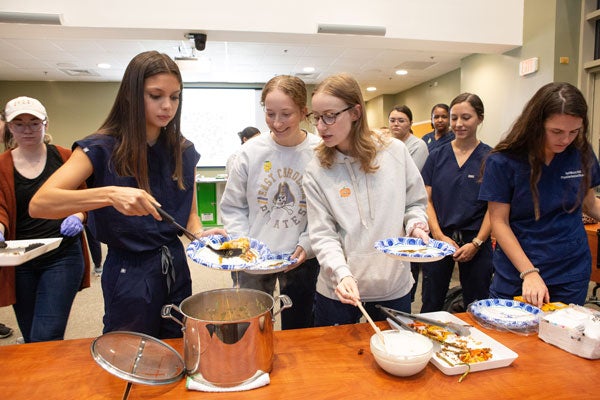 A brunette woman spoons food from a pot onto a paper plate as two women look on and others wait in line behind them.