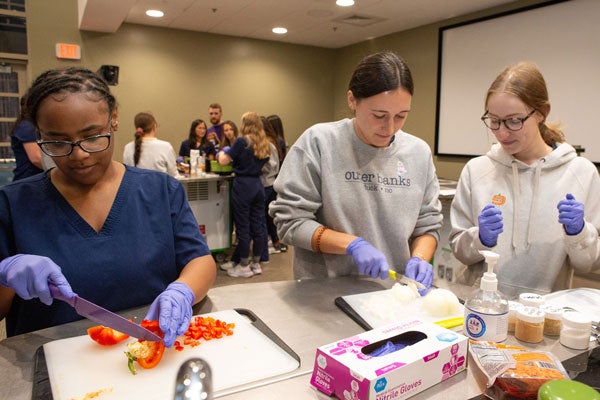 Three women with purple sterile gloves cut vegetables at a mobile kitchen prep table in a conference room with another group of people in the background working at another prep table.