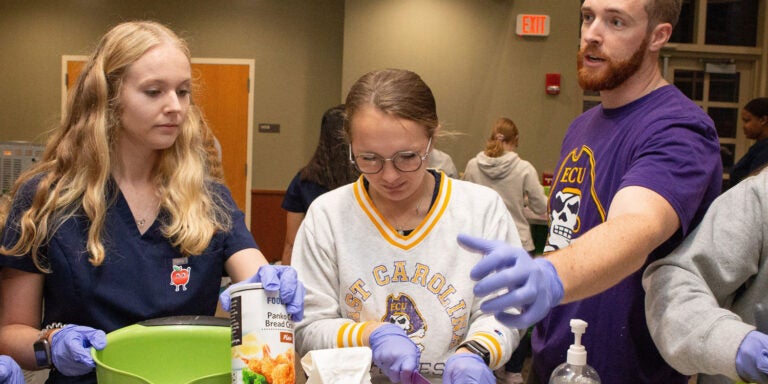 A bearded man in a purple t-shirt talks with a group of women who are preparing food in a large conference room.