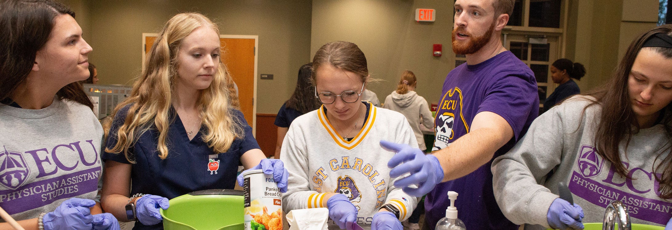 A bearded man in a purple t-shirt talks with a group of women who are preparing food in a large conference room.