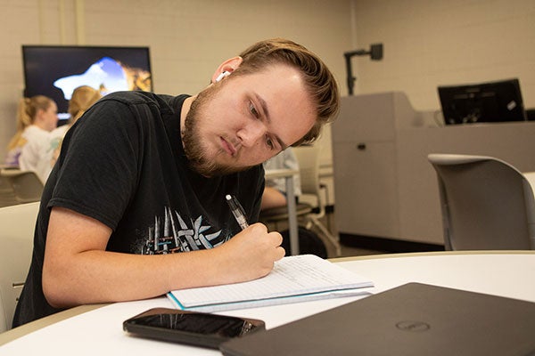 : A man in a black t-shirt writes on a piece of paper while reading information on a cell phone in a desk in a classroom.