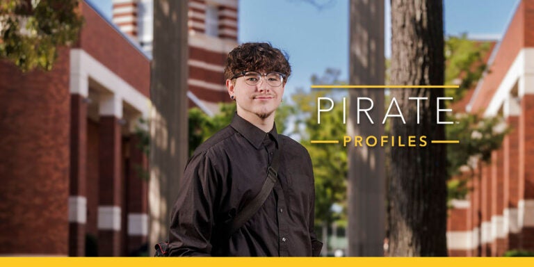 A young man with glasses wearing a black long sleeve shirt smiles and stands in front of columns and brick buildings.