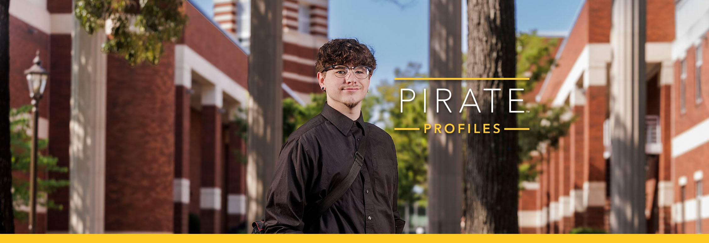 A young man with glasses wearing a black long sleeve shirt smiles and stands in front of columns and brick buildings.