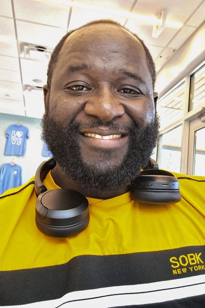 A man in a yellow shirt smiles into the camera while wearing headphones around his neck.