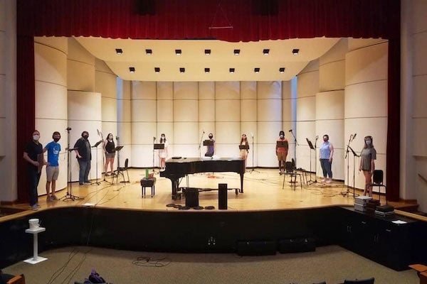 Ten people on a recital hall stage, spaced equally across, with individual microphones and music stands. They are wearing face masks. A piano stands in the middle of the stage.