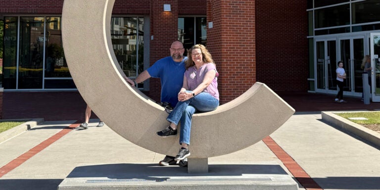 A man and woman pose outside of a brick building in the mid-day sun.