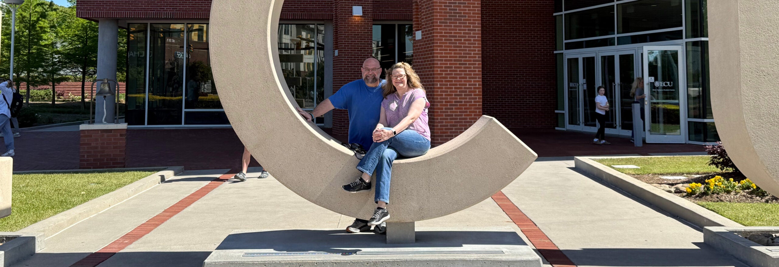 A man and woman pose outside of a brick building in the mid-day sun.