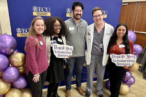 Five medical students stand before a blue photo backdrop branded ECU Brody School of Medicine – two hold small signs reading “Thank you scholarship donors.”