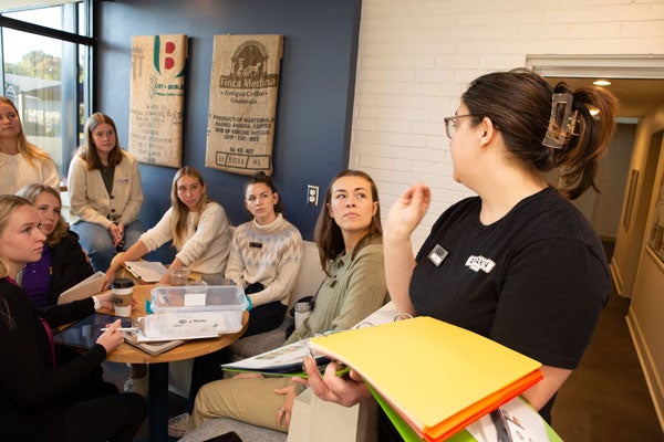 A dark-haired woman in a black T-shirt holds a binder with multi-colored paper while she speaks to a group of women in a coffee shop with white-painted brick walls.