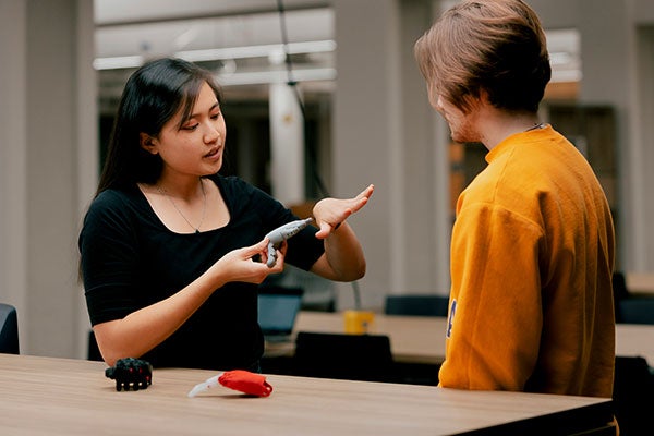 A woman demonstrates a device to a man in a casual indoor setting, with objects including a model hand on the table between them.