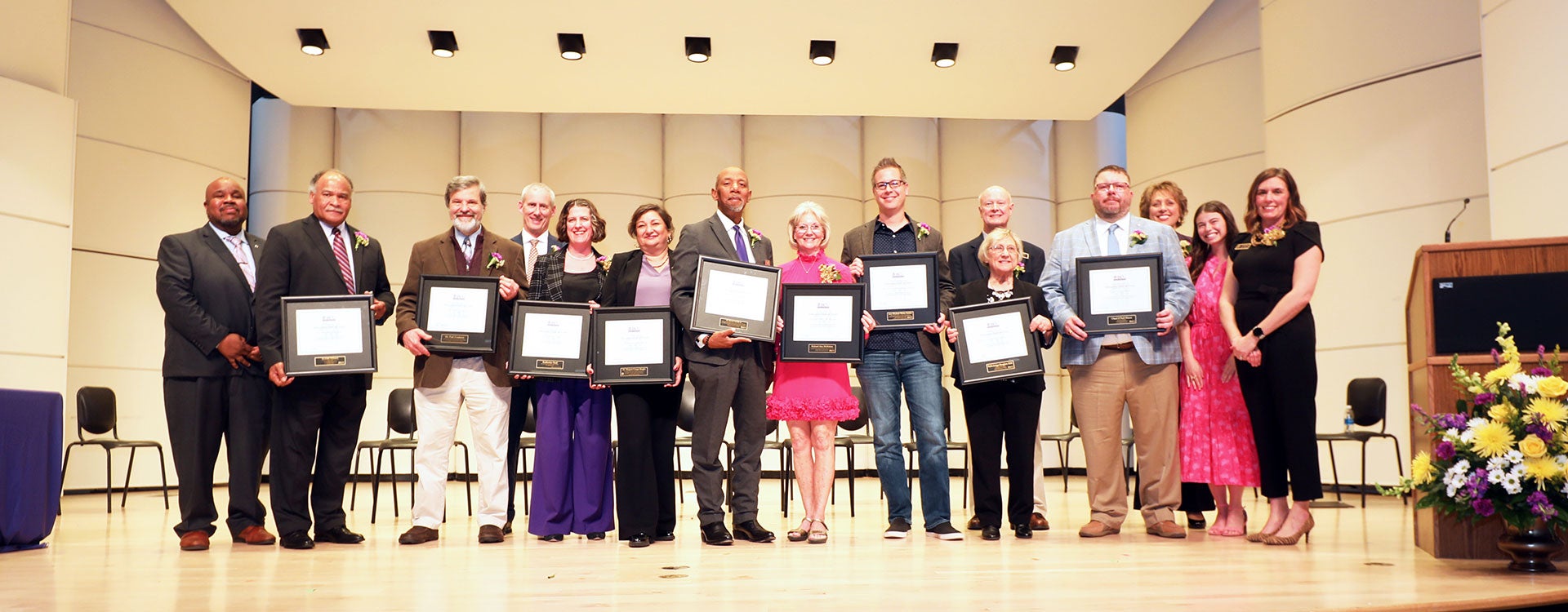 Fifteen people in business attire proudly showcase their awards while standing on a stage.