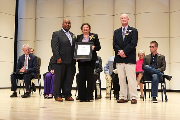 Three people stand on a stage and smile while wearing business professional attire. There is a dark-haired woman in the center holding a plaque, with two men on either side of her.