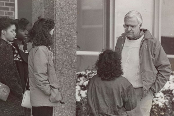 A man with white hair speaks with a group of students outside of a brick building.