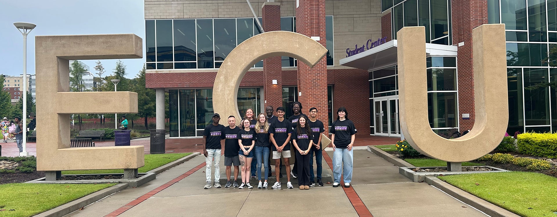 A diverse group of biomedical undergraduates and ECU faculty stand in front of the ECU letters in front of the Main Campus Student Center along 10th Street.