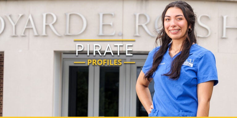 A dental student stands posing in front of Ross Hall, smiling and wearing blue scrubs.