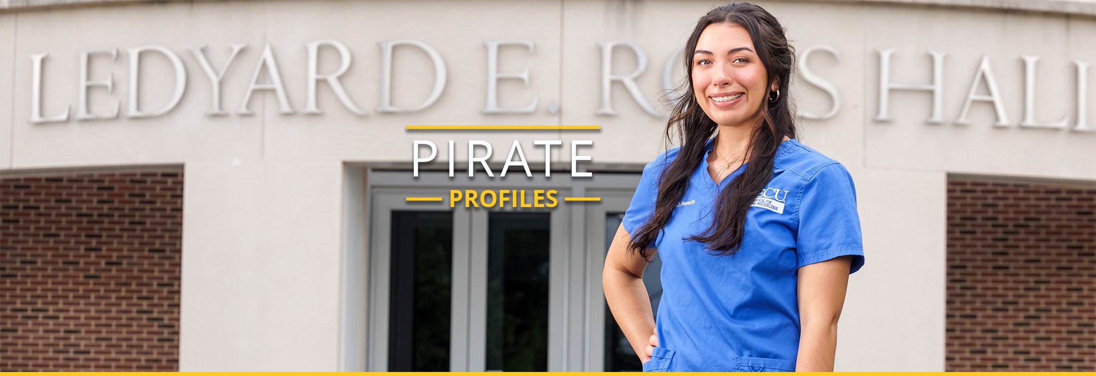 A dental student stands posing in front of Ross Hall, smiling and wearing blue scrubs.