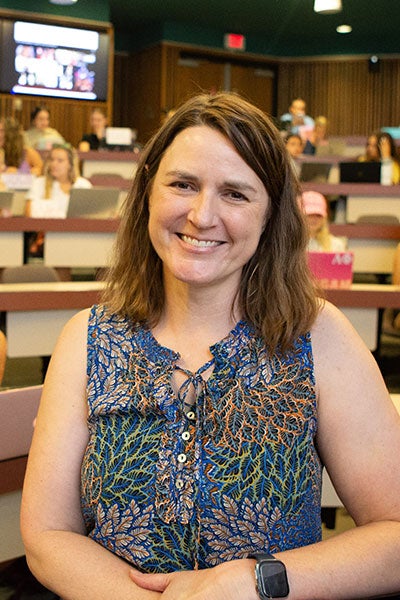 A woman with shoulder length brown hair and a floral-print top poses in front of a large stadium seating class with students seated at desks talking and working at their computers.