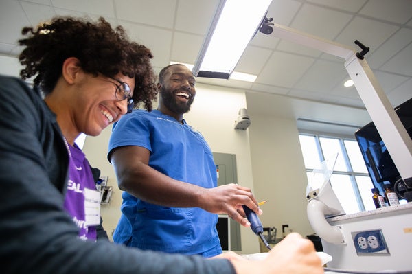 A young student, seated, is laughing while above him is an older instructor also laughing and holding a simulation dentistry tool.