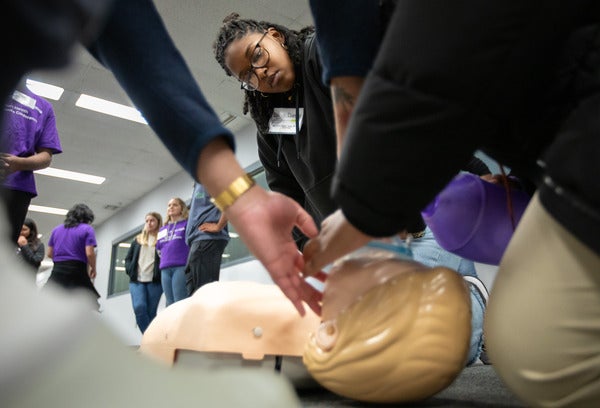 The face of a high school girl can be seen through a web of arms and hands attending to a mannequin used to execute a life support simulation. 