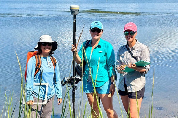 Three women wearing hats, sunglasses and boots stand in the mud of a marsh in Pine Knoll Shores, North Carolina. Each one is holding a piece of equipment or supplies for recording their observations.